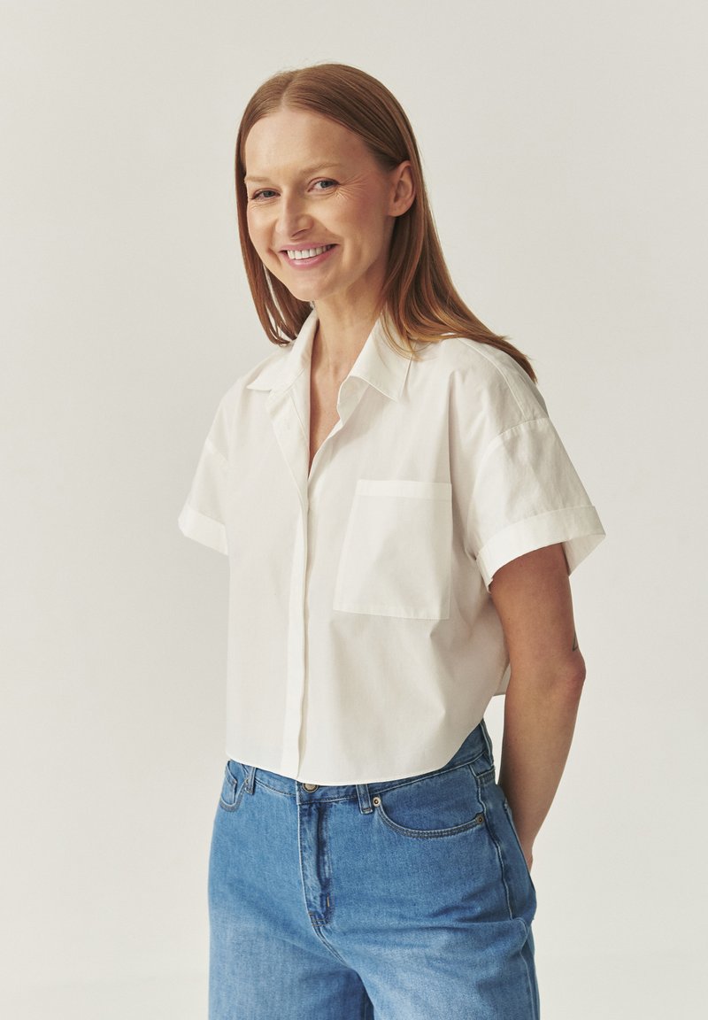 White short-sleeve button-up shirt with a pocket, made of cotton. Paired with blue denim jeans. Simple design and relaxed fit.