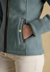 Personne portant une veste polaire bleue avec des garnitures marron, la main dans la poche latérale, et un pantalon beige sur un fond neutre.