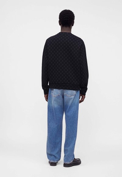Homme de dos portant un pull noir à motifs, un jean bleu ample et des chaussures noires sur un fond clair uni.