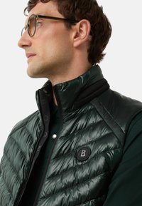 Mann mit einer dunkelgrünen Steppweste über einem dunkelgrünen Hemd, mit Brille, der nach links schaut vor einem schlichten weißen Hintergrund.