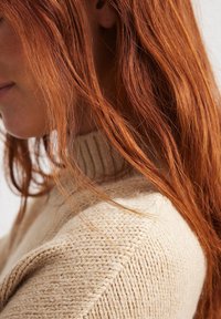 Suéter beige de punto con un cuello alto, que presenta un tejido texturizado. Larga melena roja cae sobre el hombro, ocultando parcialmente la prenda.