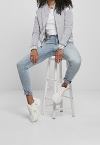 Person wearing gray bomber jacket, white shirt, light blue jeans, white sneakers, sitting on a white high stool against a plain background.