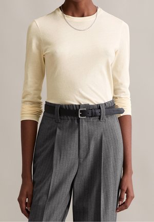 Person iført en cremefarvet skjorte med lange ærmer, stoppet ned i højttaljede grå bukser med striber, en sort bælte og et sølvfarvet kædehalskæde.