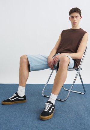 Młody mężczyzna siedzący na metalowym krzesełku składanym, ubrany w brązową bezrękawnik, jasnoniebieskie szorty z denimu, białe skarpetki i czarne trampki.