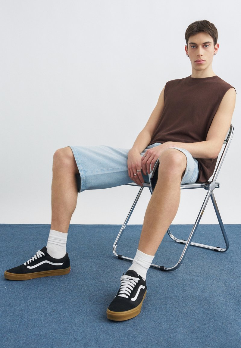 Νέος άνδρας που κάθεται σε μια μεταλλική πτυσσόμενη καρέκλα, φορόντας ένα καφέ φανελάκι χωρίς μανίκια, ανοιχτόχρωμα μπλε τζιν σορτς, λευκές κάλτσες και μαύρα αθλητικά παπούτσια.