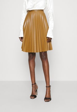 Tan plissé rok van nepleer met een aangesloten tailleband. Deze heeft verticale plooien en een uitlopende zoom, gecombineerd met zwarte hoge hakken met bandjes.