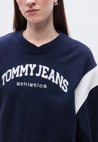 Μπλε ναυτικό φούτερ με V λαιμόκοψη, με λευκό κείμενο "TOMMY JEANS athletics" και αντίθετη λευκή διαγώνια επιφάνεια σε ένα μανίκι.