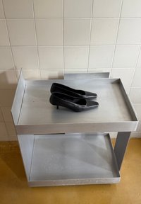 Scarpe nere con tacco alto posizionate su un tavolino in metallo argentato dal design pulito e forma rettangolare, con un ripiano inferiore.