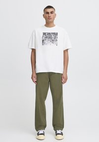 White cotton T-shirt with a black graphic print on the chest, paired with olive green relaxed-fit pants and black and white sneakers.