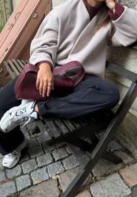 Kompaktowa bordowa skórzana torba z zaokrąglonym uchwytem, w połączeniu z szarym, oversize'owym płaszczem, ciemnymi spodniami i białymi trampkami na drewnianej ławce.