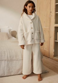 Kind steht barfuß in einer gemütlichen cremefarbenen Fleecejacke mit Holzknöpfen und passender weiter Hose in einem neutralen Schlafzimmer.