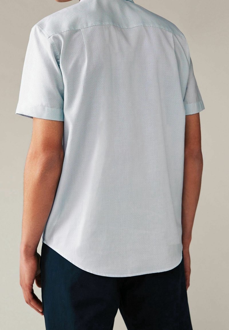 Homme portant une chemise bleu clair à manches courtes avec un motif subtil, vu de dos, accompagné d'un pantalon foncé sur fond uni.