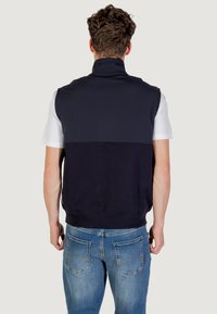 Pullover senza maniche di colore blu navy con colletto alto, realizzato in materiale testurizzato; abbinato a una maglietta bianca, indossato con jeans blu. Dettagli di cucitura visibili.