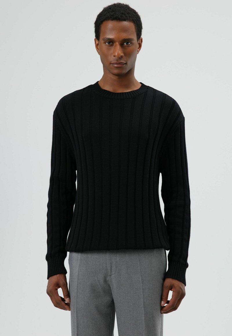 Pull noir en maille côtelée avec un col rond classique, des manches longues et une finition texturée, associé à un pantalon gris clair.