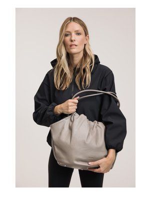 Blond vrouw met zwart jasje houdt een grote zachte grijze handtas met dubbele handvatten vast tegen een effen achtergrond.