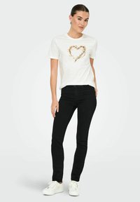 Camiseta blanca con diseño de corazón dorado, combinada con jeans negros y zapatillas blancas. La camiseta tiene mangas cortas y un escote redondo.
