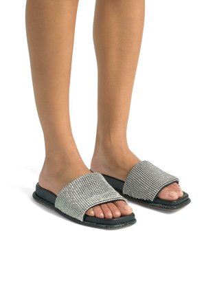 Silver rhinestone-embellished slide sandals with a black rubber sole and flat design, revealing toes and smooth foot texture.