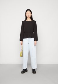 Top preto de manga longa com cintura franzida; jeans de perna larga azul claro; segurando uma pequena mala verde; sapatos pretos robustos.