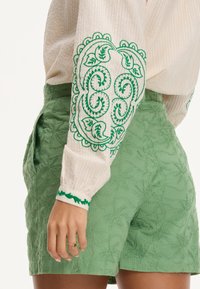 Blusa bianca texturizzata con ricami verdi sulle maniche, abbinata a pantaloni corti verdi con un motivo a foglie. Anello semplice visibile.