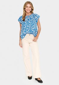 Top floreale blu con maniche corte, dettaglio a chiave, abbinato a pantaloni beige a campana e ballerine nere. Tessuto liscio con una vestibilità comoda.