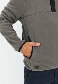 Grå fleecepullover med en sort knaplukning, kontrasterende sort kant, sidelommer og et lille logo-patch på den nederste kant.