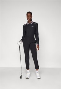 Svart golfoutfit som består av en långärmad polo med rosa och vita ränder, slim fit-byxor och vita golfskor, håller en golfklubba.