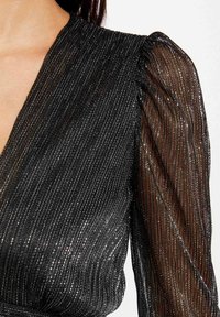 Zwarte jurk met lange, doorzichtige pofmouwen en een geribbelde textuur. De stof bevat metallic draden voor een glanzend effect.