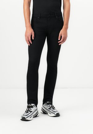 Person wearing fitted black pants and silver, black, and white sneakers, standing against a plain white background.
