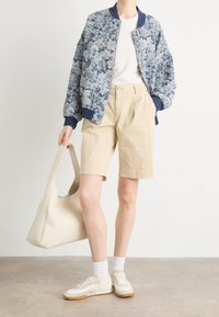 Giacca bomber floreale blu con polsini a coste, t-shirt bianca, pantaloncini beige, sneakers bianche con suola beige e borsa a spalla crema.