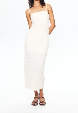 Kobieta ubrana w białą, bezrękawnikową, teksturowaną sukienkę maxi z cienkimi ramiączkami i czarne sandały na obcasie z paskami, stoi z rękami za plecami.