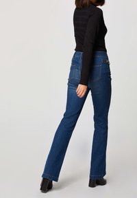 Modré džíny s rozšířenými nohavicemi a středním pasem, s dvěma zadními kapsami a detailem knoflíku na poutku na opasek. Modelka má na sobě černé tričko s dlouhým rukávem.