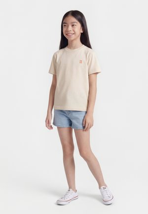 Mosolygó, hosszú hajú lány bézs pólóban narancssárga logóval, világoskék farmer rövidnadrágban és fehér tornacipőben, fehér háttér előtt áll.