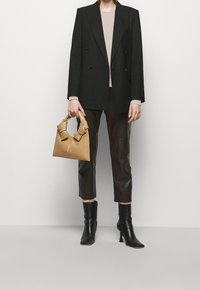 Blazer nero doppiopetto sopra un top beige, pantaloni in pelle marrone. Tiene una borsa color cammello con dettaglio a fiocco. Stivaletti neri con tacco.