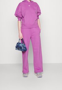 Pink hoodie with dropped shoulders and a front pocket, paired with matching wide-leg pants. Holds a blue patterned handbag. Gray sneakers.