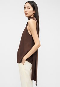 Καφέ αμάνικο τοπ με ψηλό λαιμό και ρέον πίσω μέρος, κατασκευασμένο από λείο ύφασμα. Συνδυάζεται με ανοιχτόχρωμο παντελόνι.