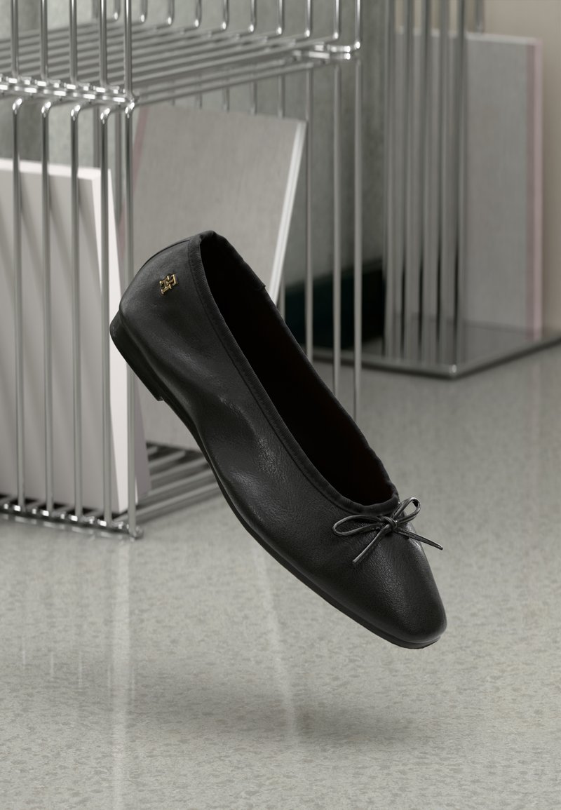 Ballerine en cuir noir avec petit nœud et logo doré, positionnée en suspension au-dessus d'un sol tacheté et d'un porte-livres en fil métallique.