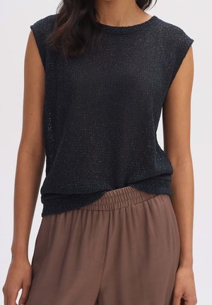 Femme portant un haut en maille noir sans manches, rentré dans un pantalon marron ample avec une taille élastique, visible des épaules aux hanches.