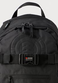 Zaino nero realizzato in tessuto resistente, dotato di tasche con doppia cerniera, una tracolla per skateboard ripiegabile e un logo testurizzato sulla parte frontale.