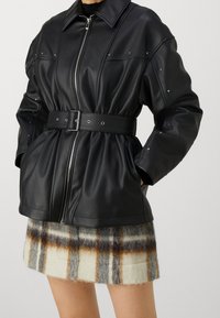 Giubbotto in pelle sintetica nera con vita cinturata, chiusura zip e borchie decorative sulle maniche. Indossato sopra una mini gonna in lana a quadri.