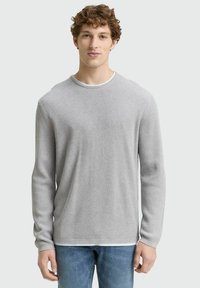 Grauer gerippter Pullover mit langen Ärmeln, Rundhalsausschnitt und einem weißen Lagen-Detail am Kragen. Getragen mit hellblauen Jeans.