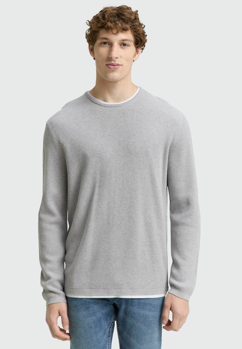 Grauer gerippter Pullover mit langen Ärmeln, Rundhalsausschnitt und einem weißen Lagen-Detail am Kragen. Getragen mit hellblauen Jeans.