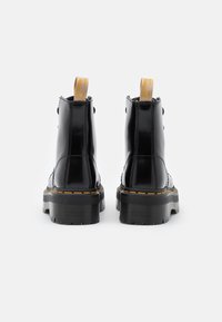 Mustan kiiltonahkakengät nilkkaan, joissa on paksu pohja, keltainen vetotappi ja erottuva keltainen tikkaus pohjassa ja sauman varrella.