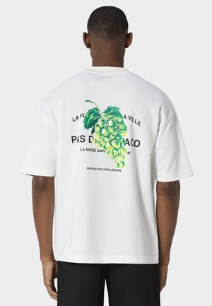 Uomo che indossa una t-shirt bianca con una vite verde e testo francese stampato sulla schiena, abbinata a pantaloni neri.