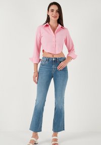 Pink cropped button-up shirt with long sleeves, paired with light blue flared jeans. Model wears white heeled sandals. Simple, modern design.