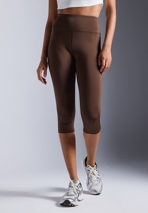 Brown, form-fitting capri leggings with a high waistband, made of smooth material. White athletic shoes with silver accents are worn.
