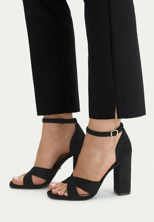 Pies con sandalias negras de tacón alto con tiras al tobillo y tacones anchos, combinadas con pantalones negros recortados con aberturas laterales.