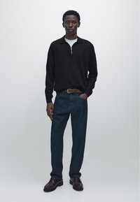Young man standing against a plain background wearing a black zip-up sweater, white undershirt, dark jeans, brown belt, and dark dress shoes.