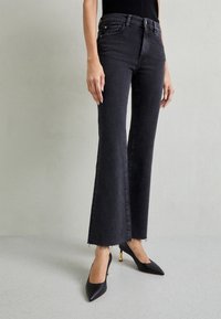 Jeans negros acampanados con dobladillo sin acabar y bolsillos delanteros, combinados con tacones negros de punta afilada con acentos en tono dorado. Textura de denim suave.