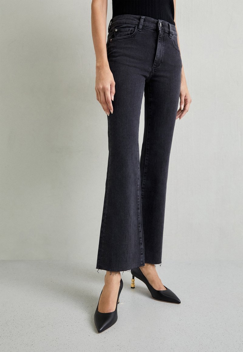 Jeans negros acampanados con dobladillo sin acabar y bolsillos delanteros, combinados con tacones negros de punta afilada con acentos en tono dorado. Textura de denim suave.