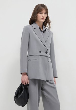 Woman wearing a light gray double-breasted blazer and matching trousers, holding a black soft leather handbag against a plain background.